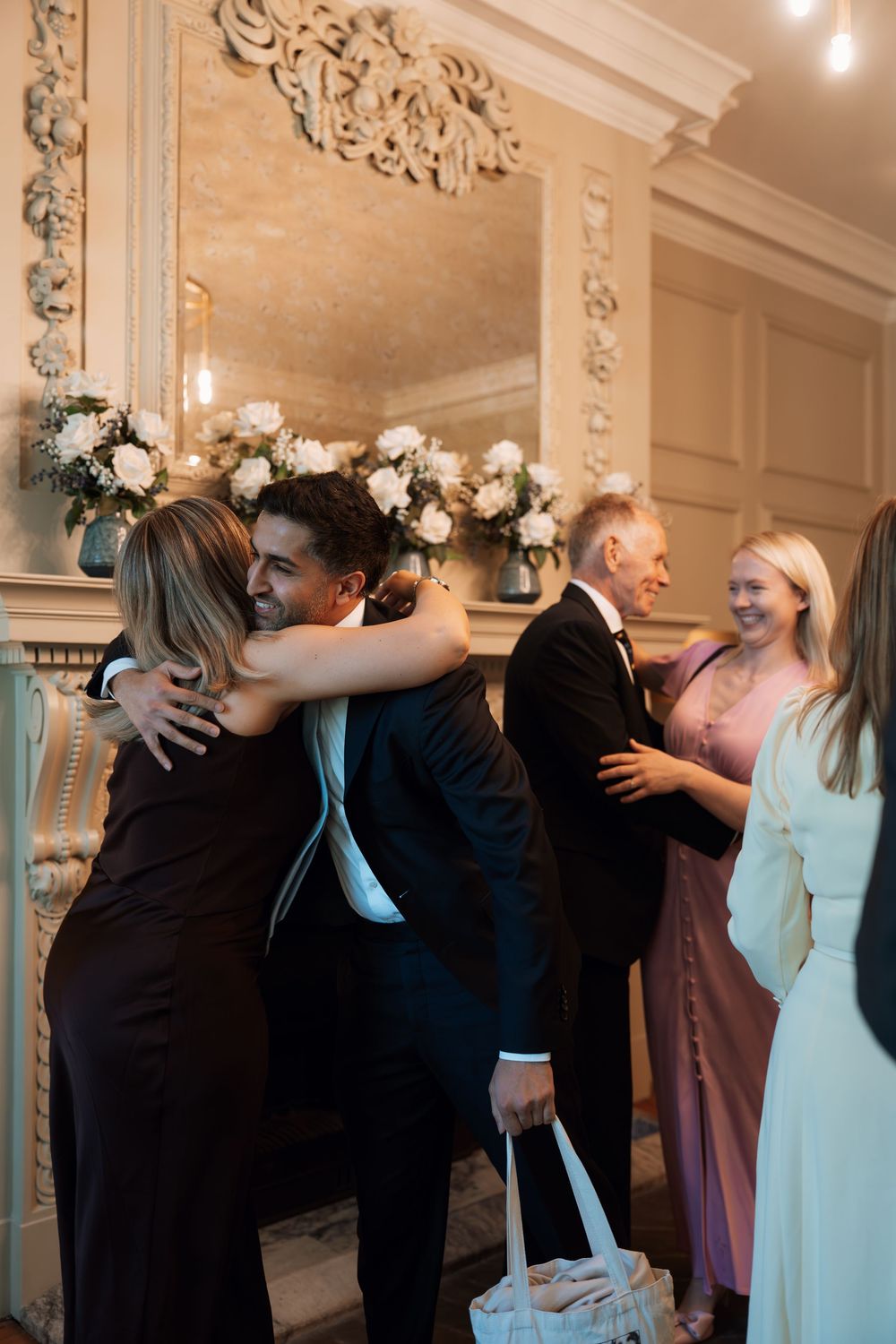 Guests in formal attire hugging and greeting each other at an elegant wedding reception near a decorative fireplace.
