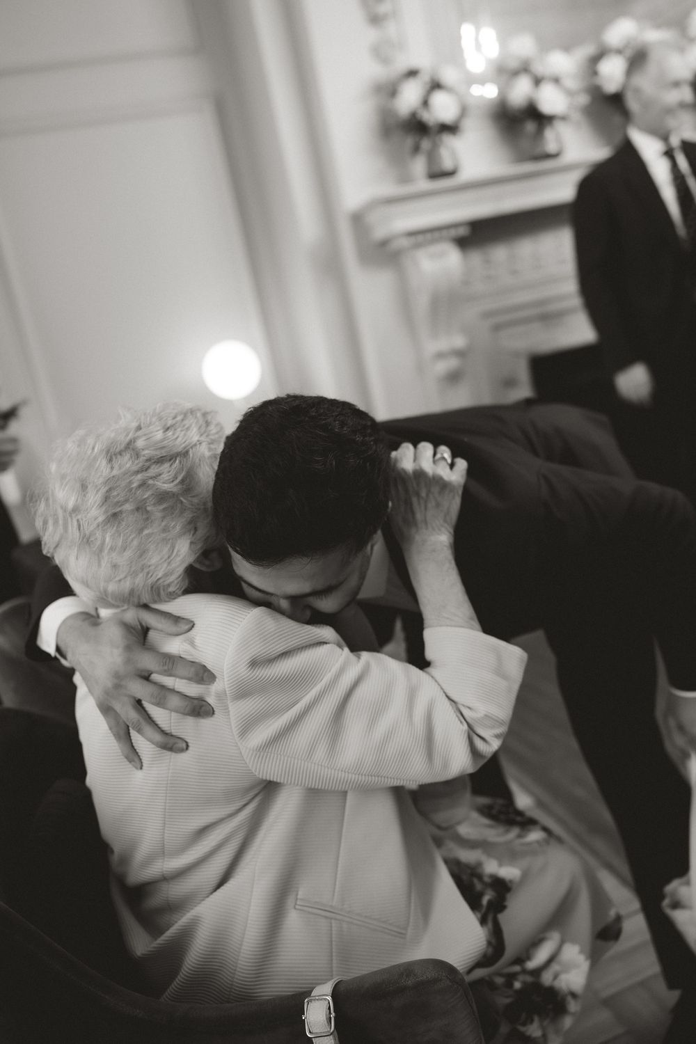 Black and white photo of a heartfelt hug between two people at a formal wedding ceremony indoors.
