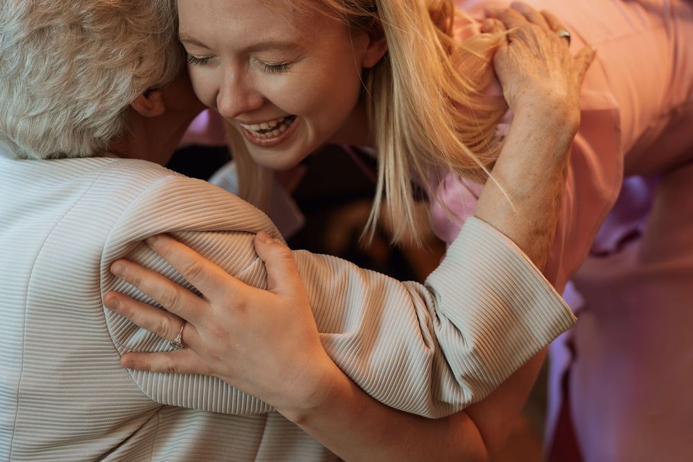 Women sharing a warm group hug, smiling joyfully in soft pink tones, capturing a heartfelt moment of friendship.