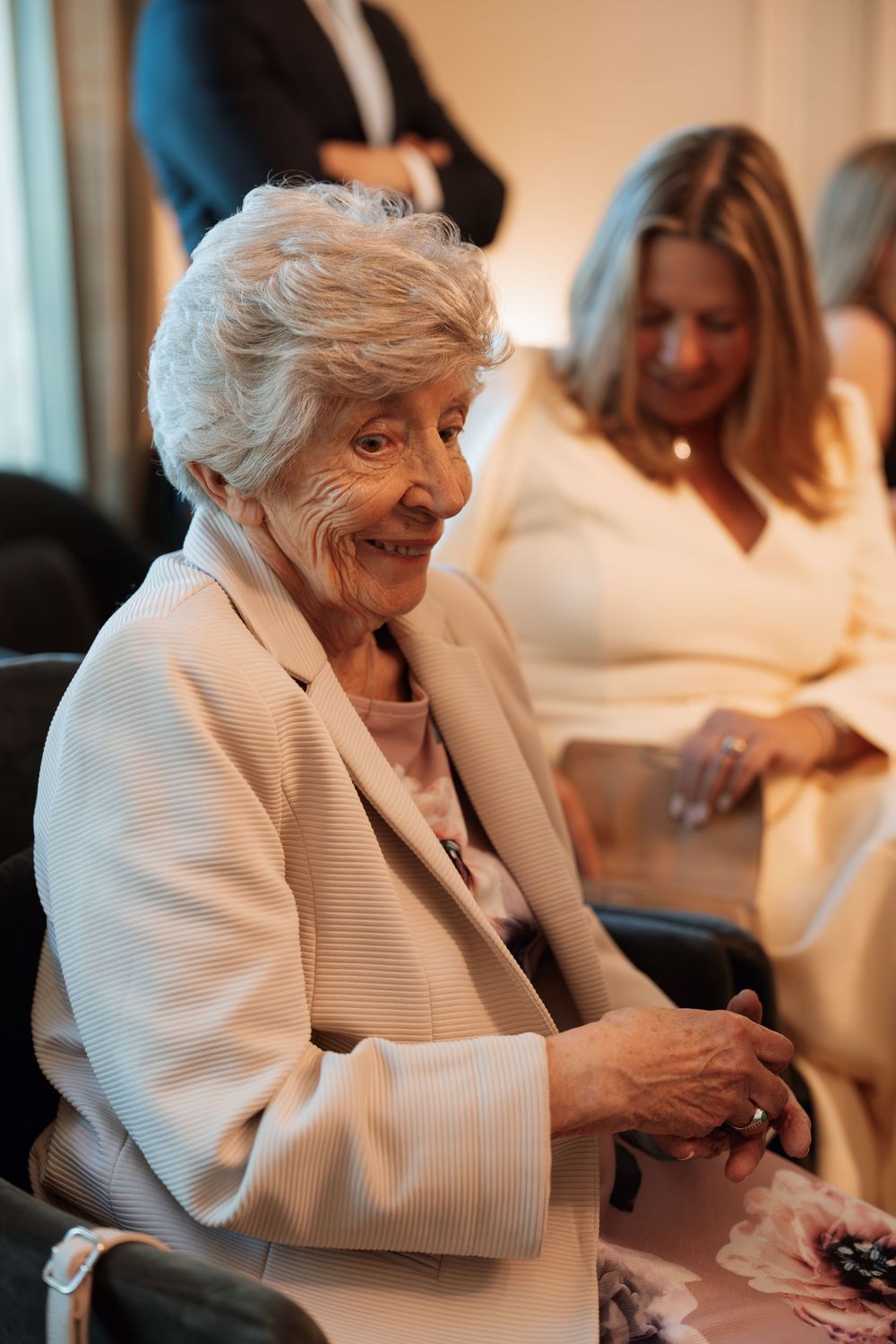 Elderly woman in white blazer smiling warmly at a formal event, with guests seated in background.