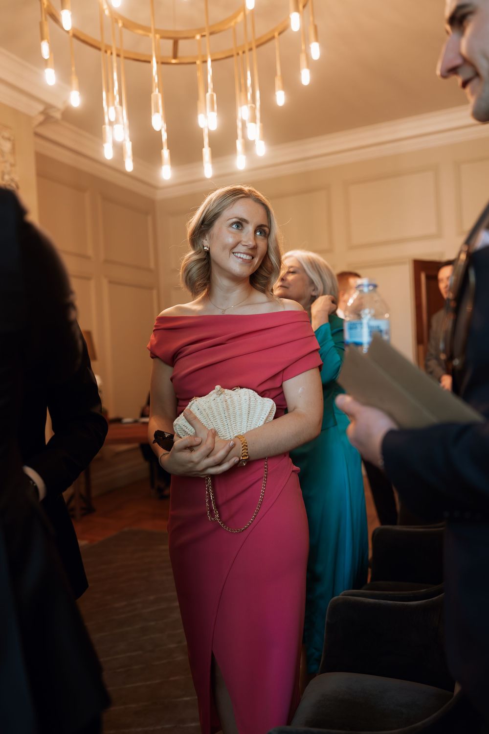 Elegant woman in pink off-shoulder gown holding shell clutch at formal event under warm chandelier lighting.