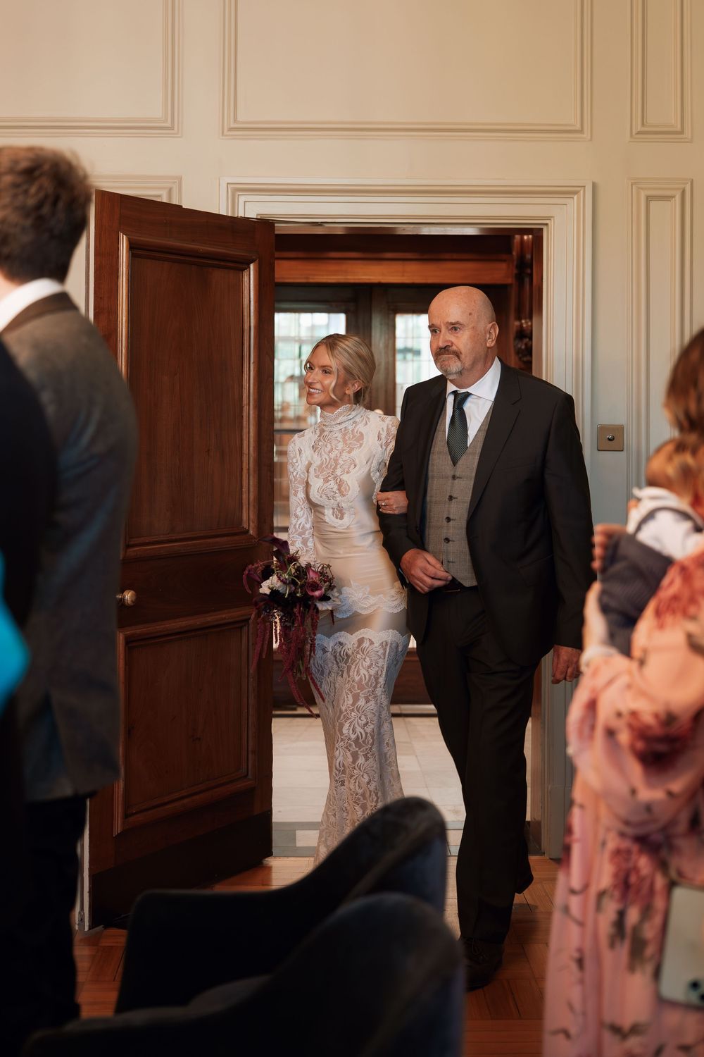 Bride in lace gown walks down the aisle with escort through elegant wood-paneled doorway at indoor wedding ceremony.