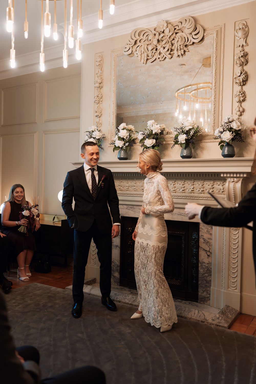 Bride in lace gown and groom in dark suit exchange vows before ornate fireplace with white floral arrangements.
