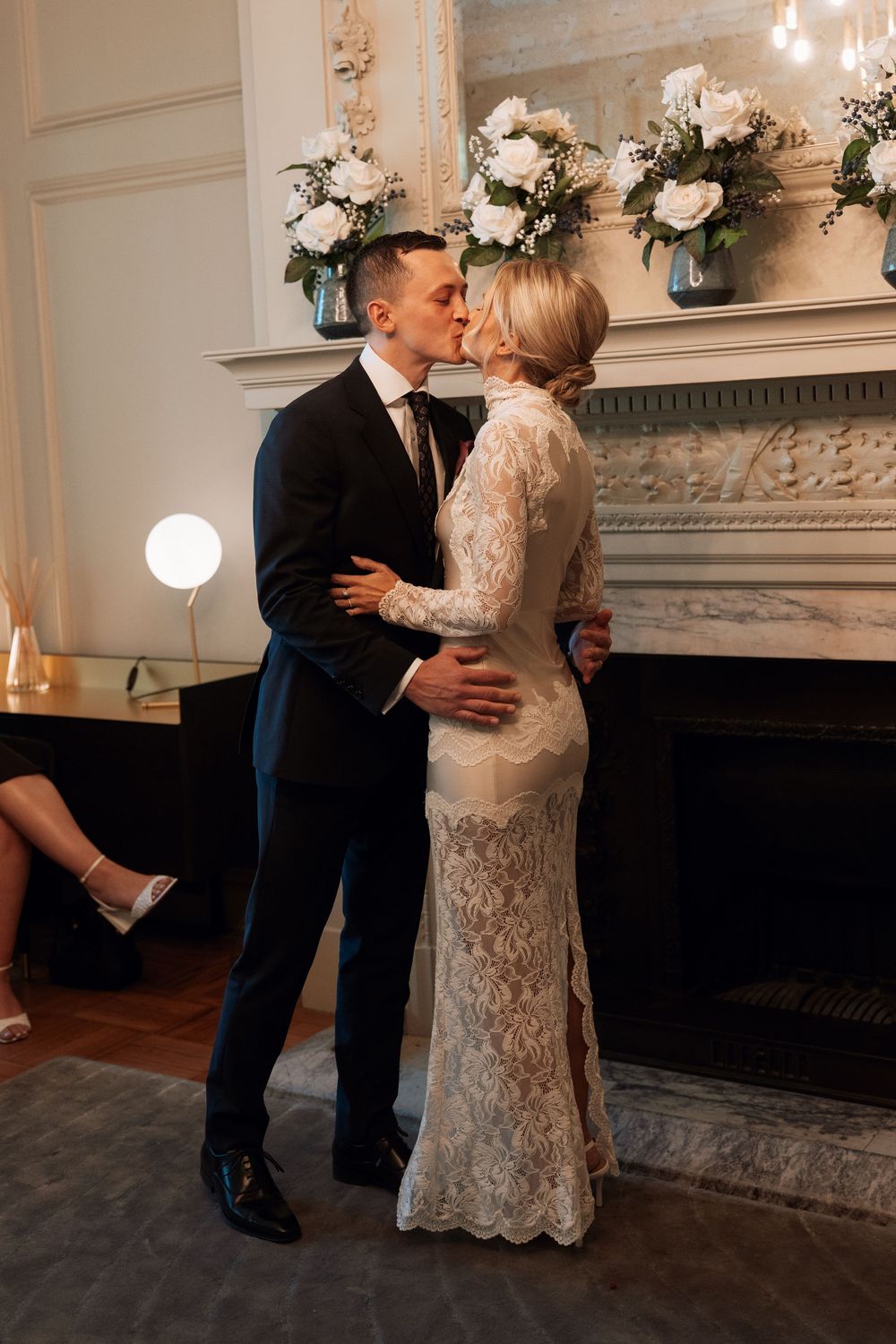 Bride and groom sharing a kiss in front of an elegant fireplace adorned with white roses during their wedding ceremony.