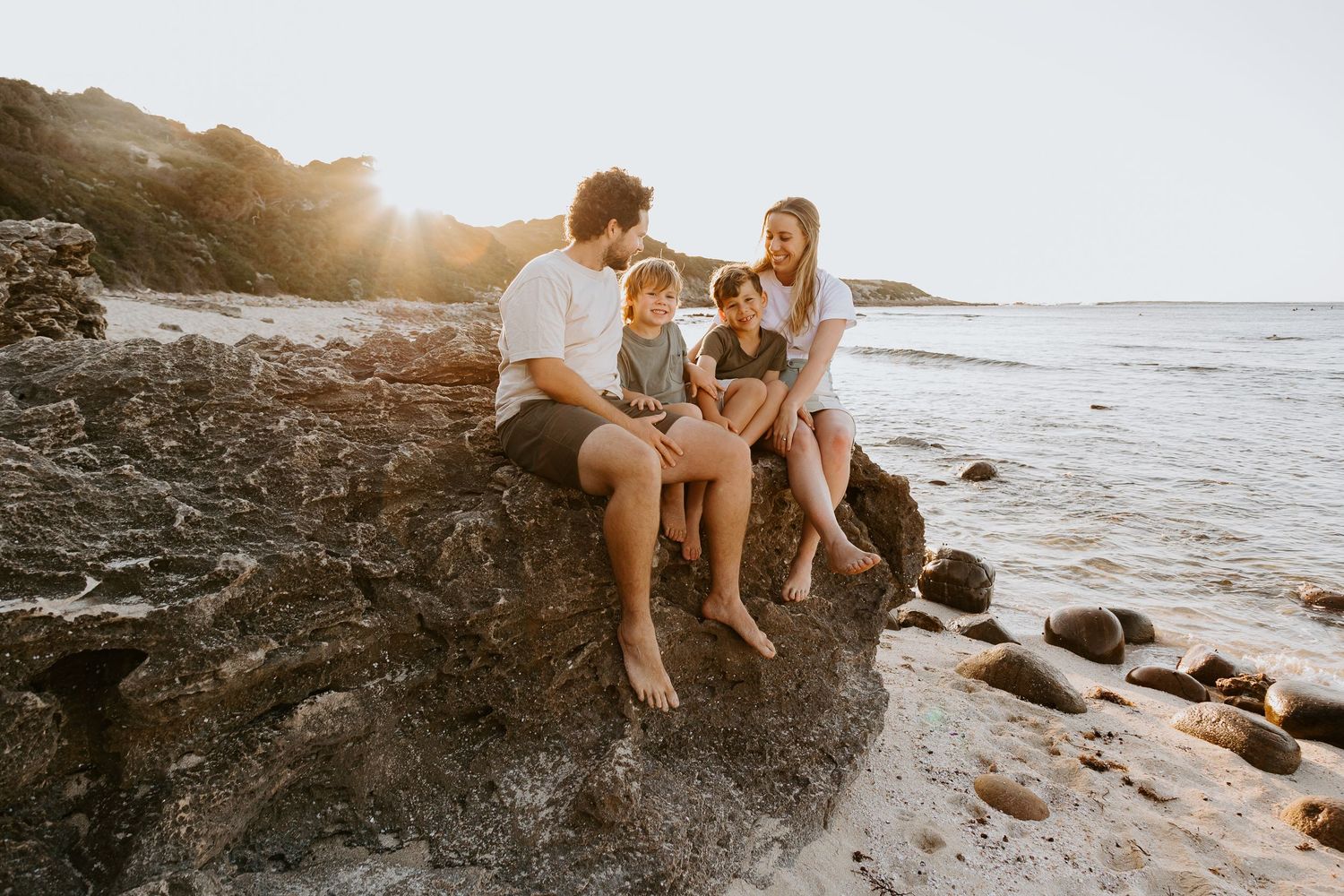 Family of four sitting on rocky beach at golden sunset with lush green hillside backdrop.
