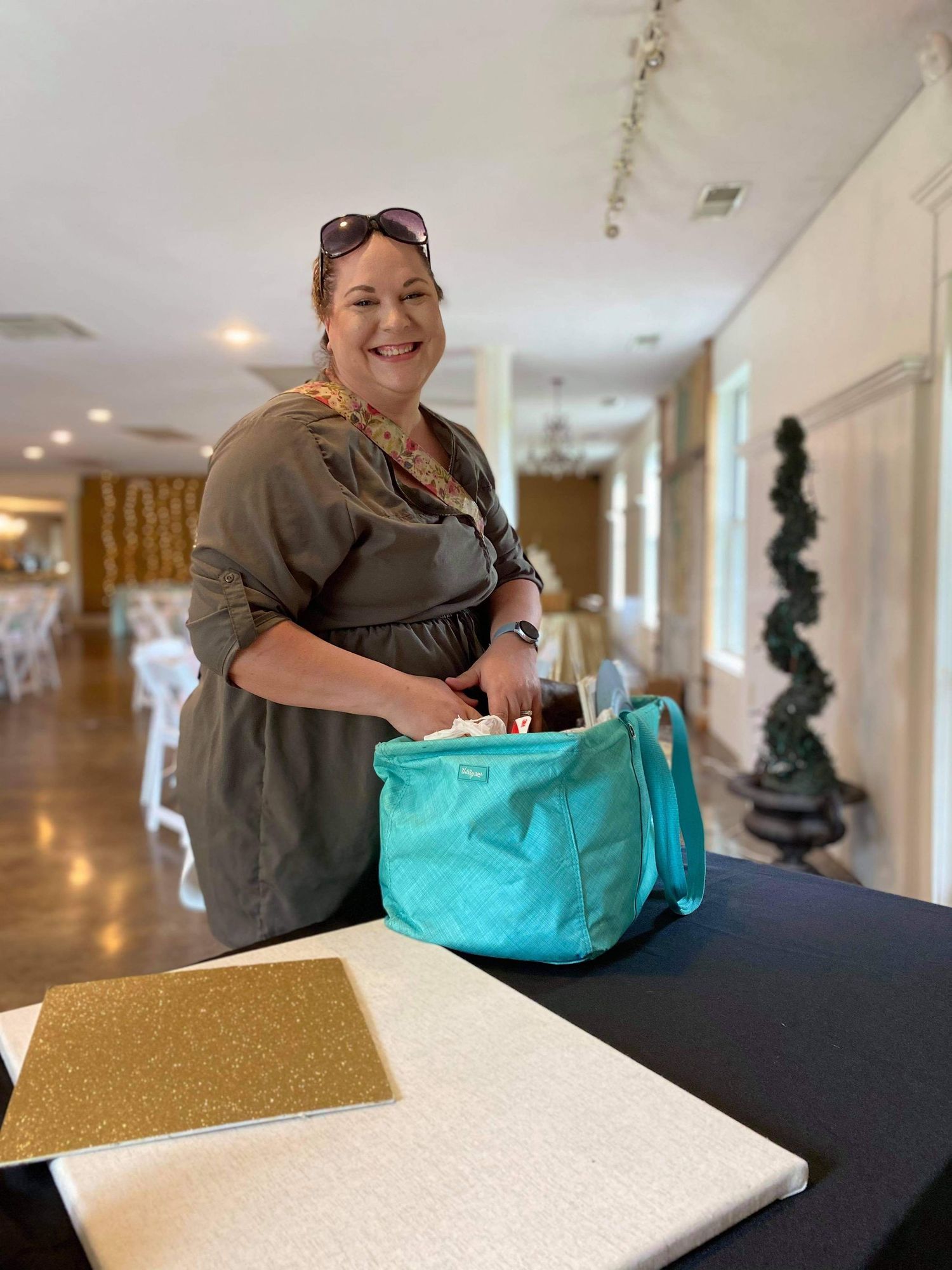 Smiling woman in olive dress holding teal bag at black table in elegant event venue.