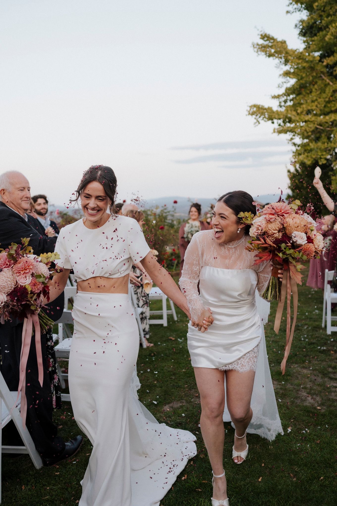 Two brides walk hand-in-hand down the aisle, smiling joyfully, holding colorful bouquets at an outdoor wedding ceremony.