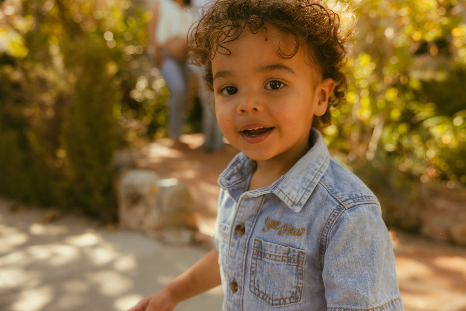 Adorable toddler in denim shirt smiling outdoors in sunny garden setting.