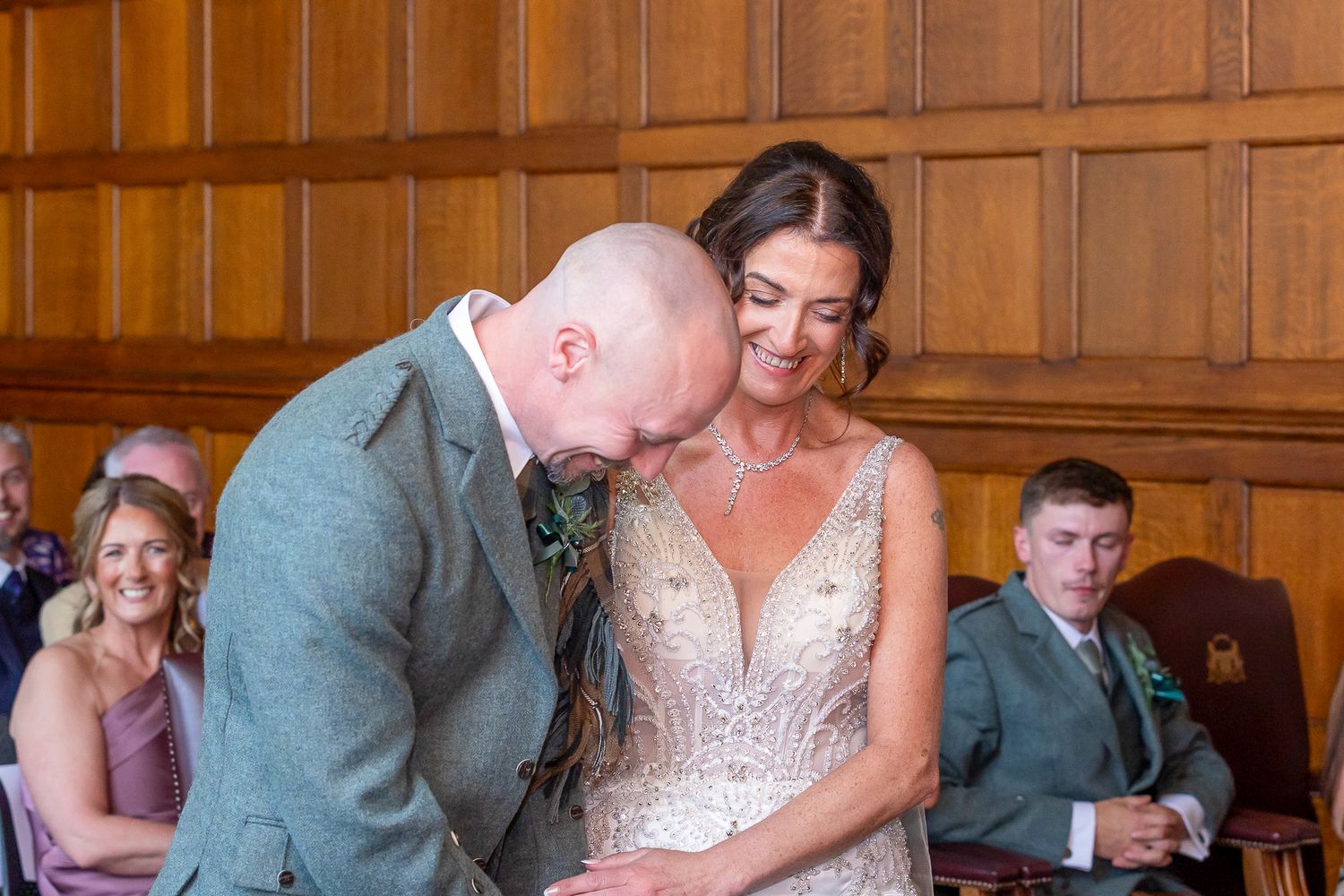 Groom kissing bride on cheek during wedding ceremony in elegant wood-paneled room with guests seated behind.