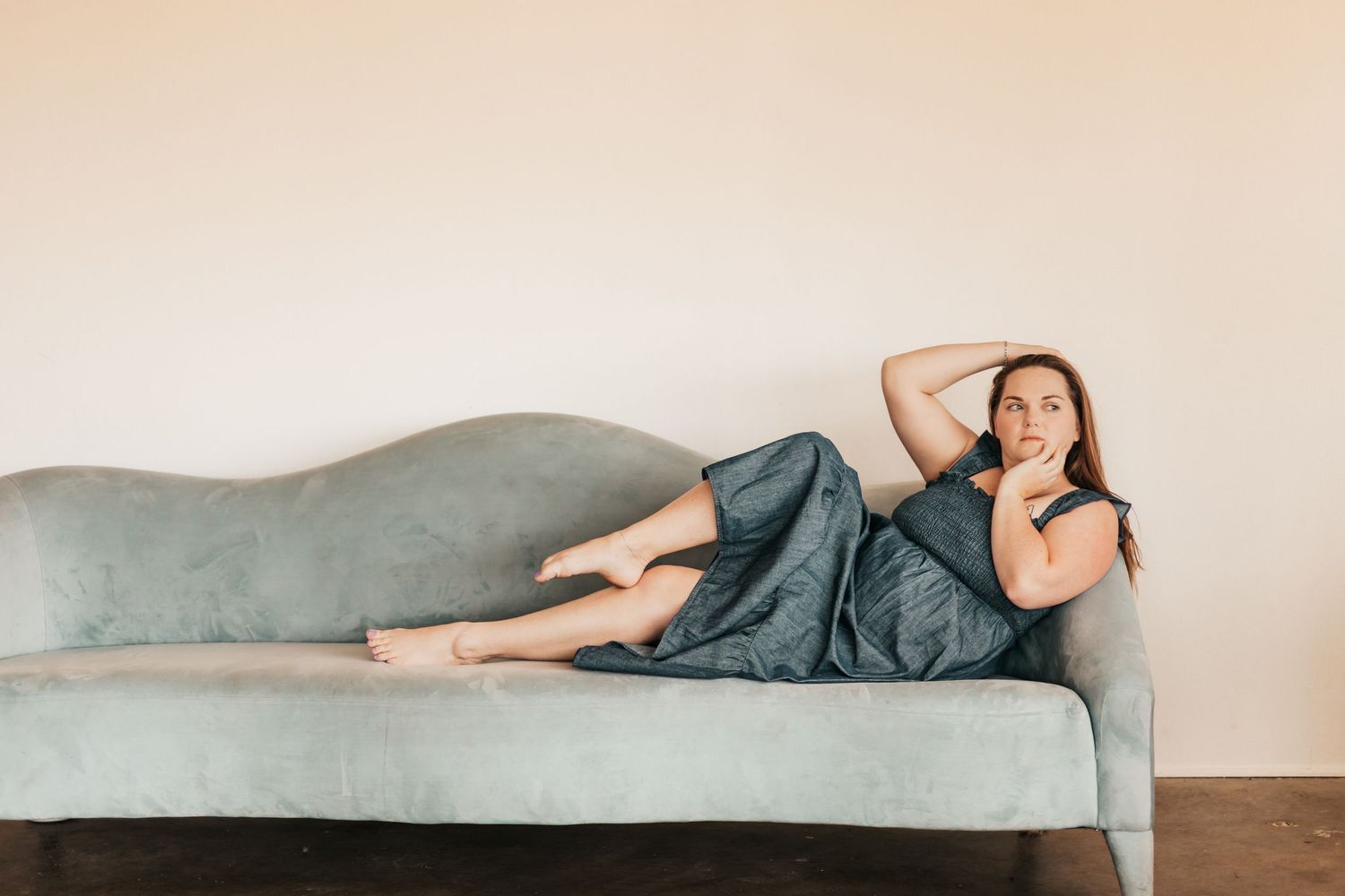 Woman in gray dress reclining on a light blue velvet sofa, gazing thoughtfully in a minimalist room | Photography Business Coach
