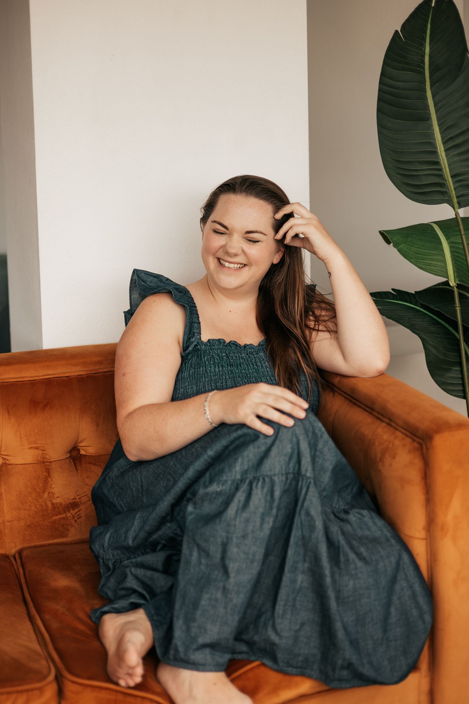 Smiling woman in teal dress sitting on brown leather sofa with tropical plant in background | Photography Business Coach