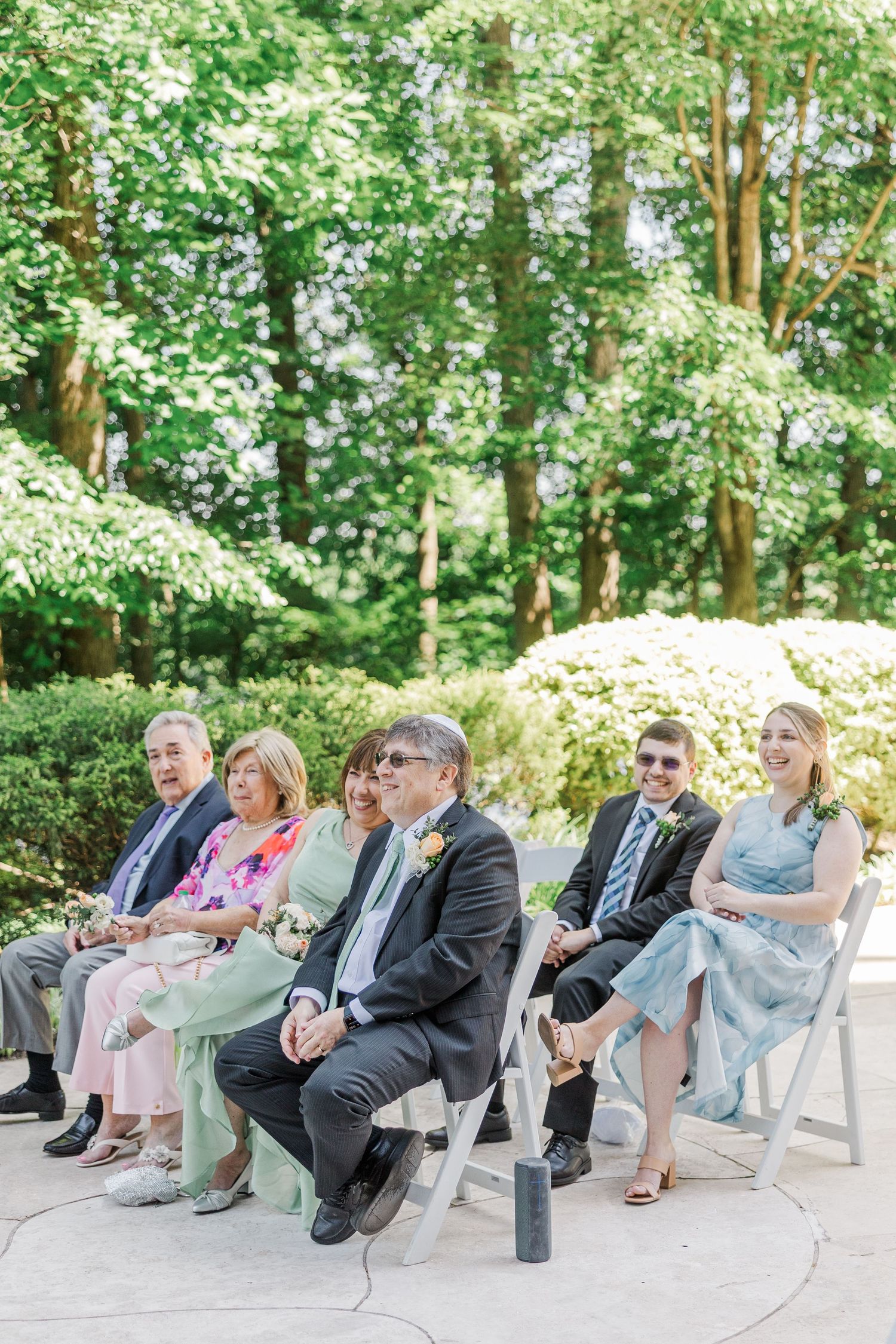 Guests seated on white chairs at an outdoor wedding ceremony surrounded by lush green trees and blooming shrubs.