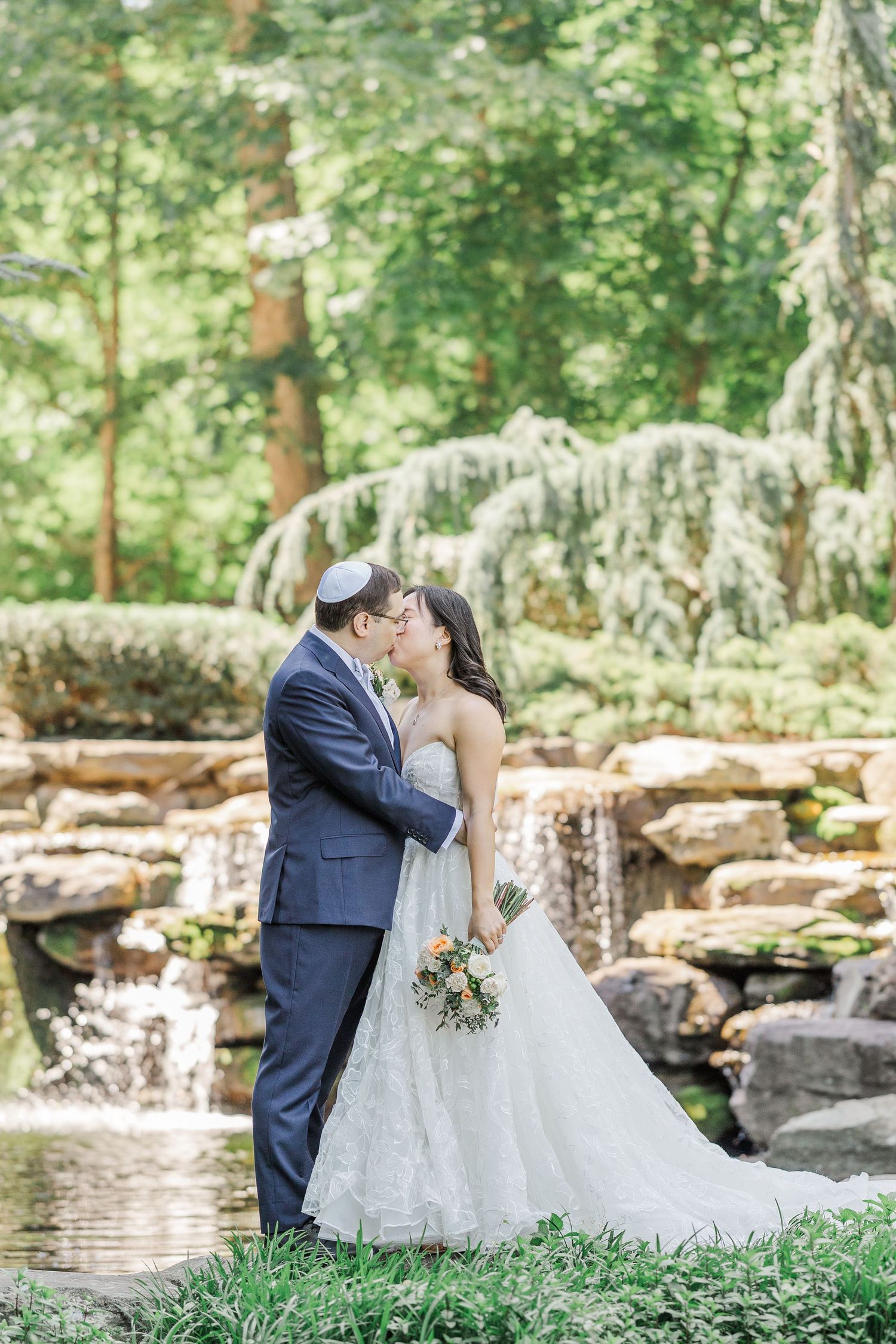 Bride and groom share a kiss in front of a stone waterfall surrounded by lush greenery at an outdoor wedding.