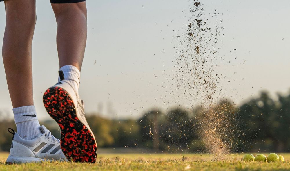 Golfer's foot in Adidas cleats kicking up dirt and grass on a golf course with tennis balls nearby.