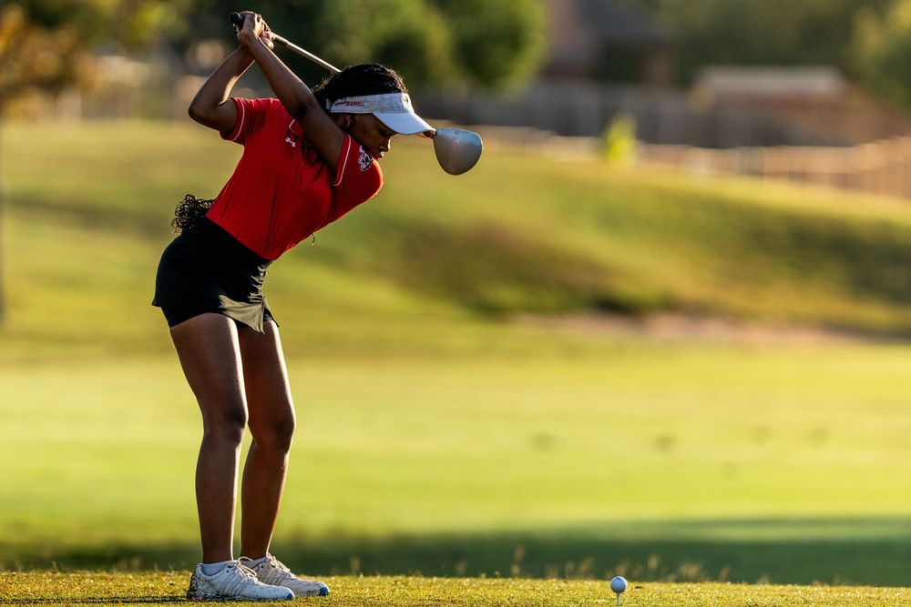 Female golfer in red shirt and visor swings a driver on a sunny golf course.
