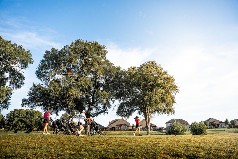 Golfers walking with bags near large oak trees on a sunny golf course.