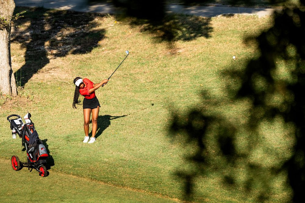 Female golfer in red shirt chipping a shot on a tree-lined golf course.