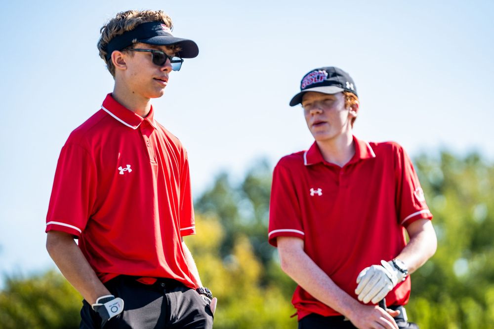 Two young male golfers in red Under Armour shirts talking on a golf course.
