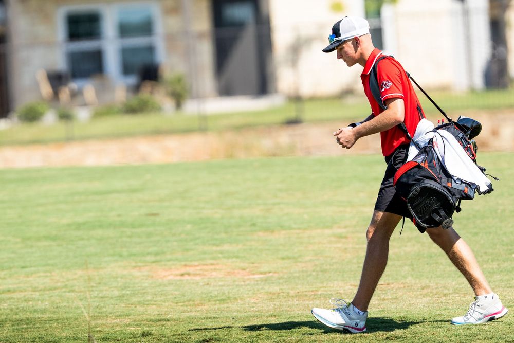 Male golfer in red shirt carrying a golf bag across a sunny fairway.