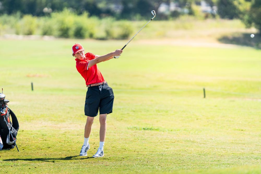 Young golfer in red shirt executes a follow-through swing on a sunny golf course.