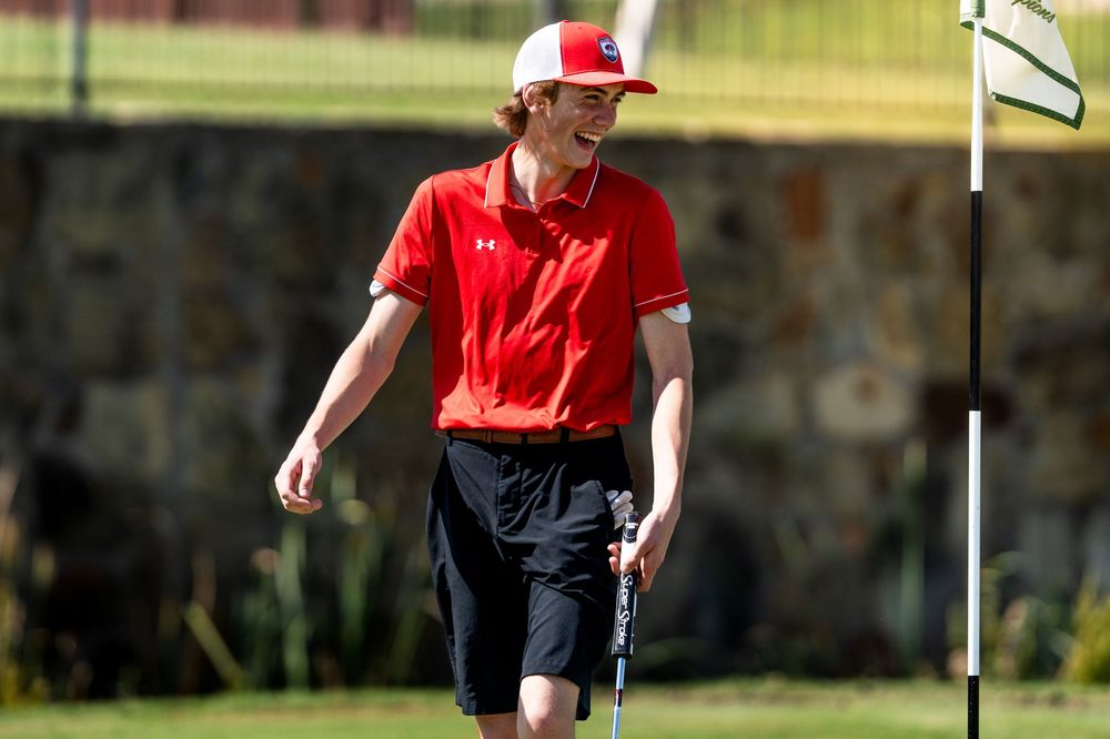 Smiling young golfer in red polo and cap stands near flag on golf course green.