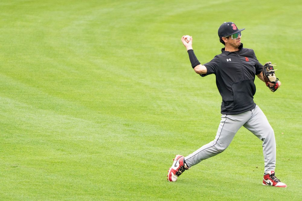 Baseball player in red and black uniform throws ball across a green outfield.