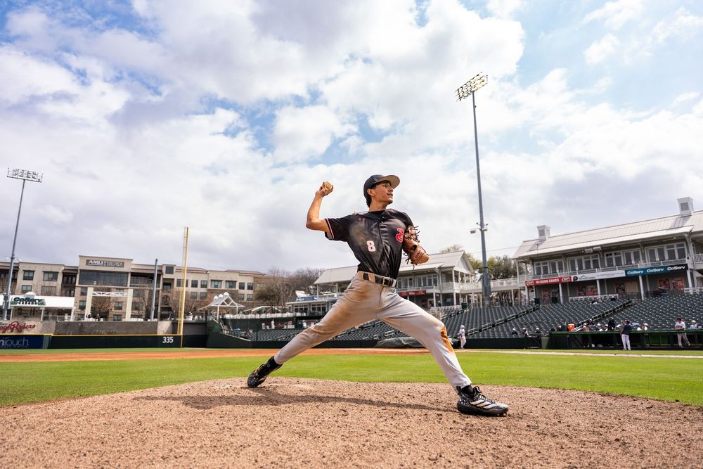 Baseball pitcher in dark uniform delivers a pitch at a professional stadium under cloudy skies.