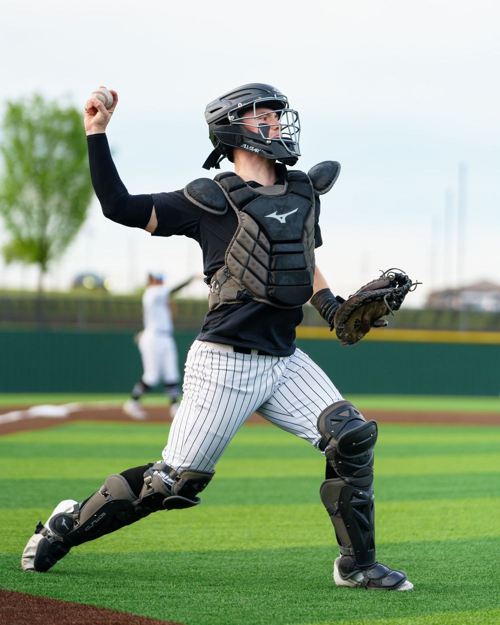 Baseball catcher in full Mizuno gear throws the ball during a game on a green turf field.