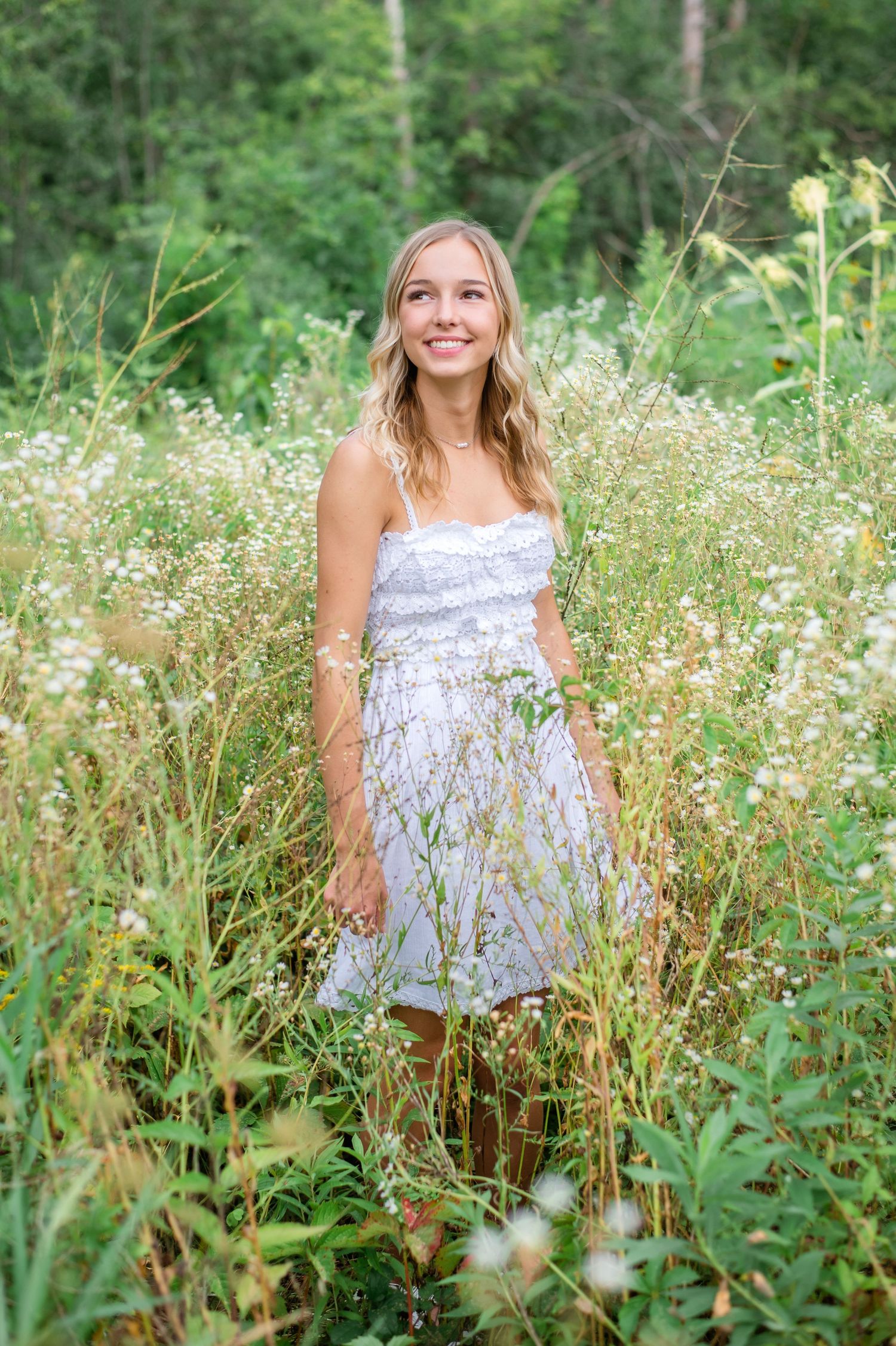 Smiling young blonde woman in white sundress standing among wildflowers and tall grass in a lush green field.