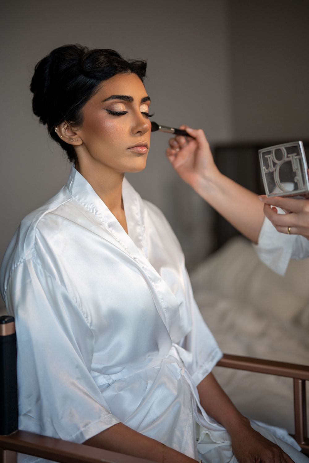 A bride in a white satin robe gets her eye makeup applied by a professional artist before her wedding.