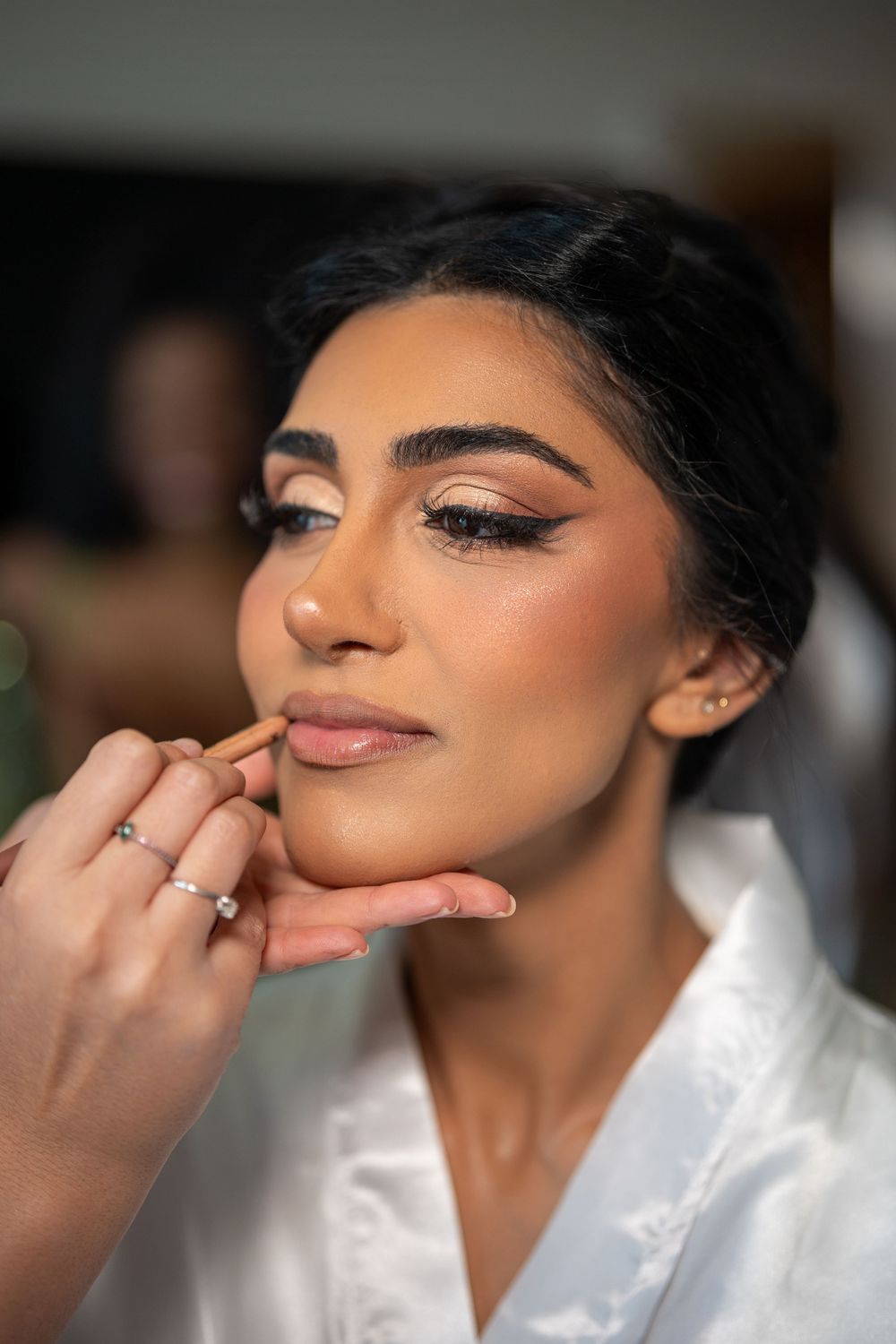 Bride getting makeup applied with elegant cat-eye liner and glowing skin, wearing white satin robe.