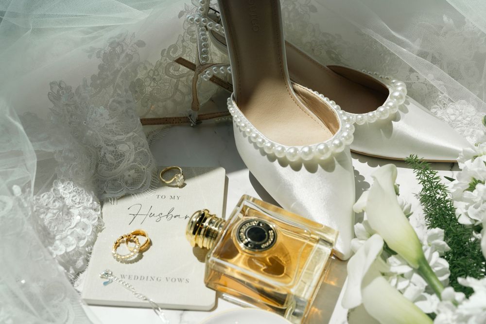 Elegant bridal flat lay with pearl heels, gold perfume bottle, wedding rings, and lace veil on white background.