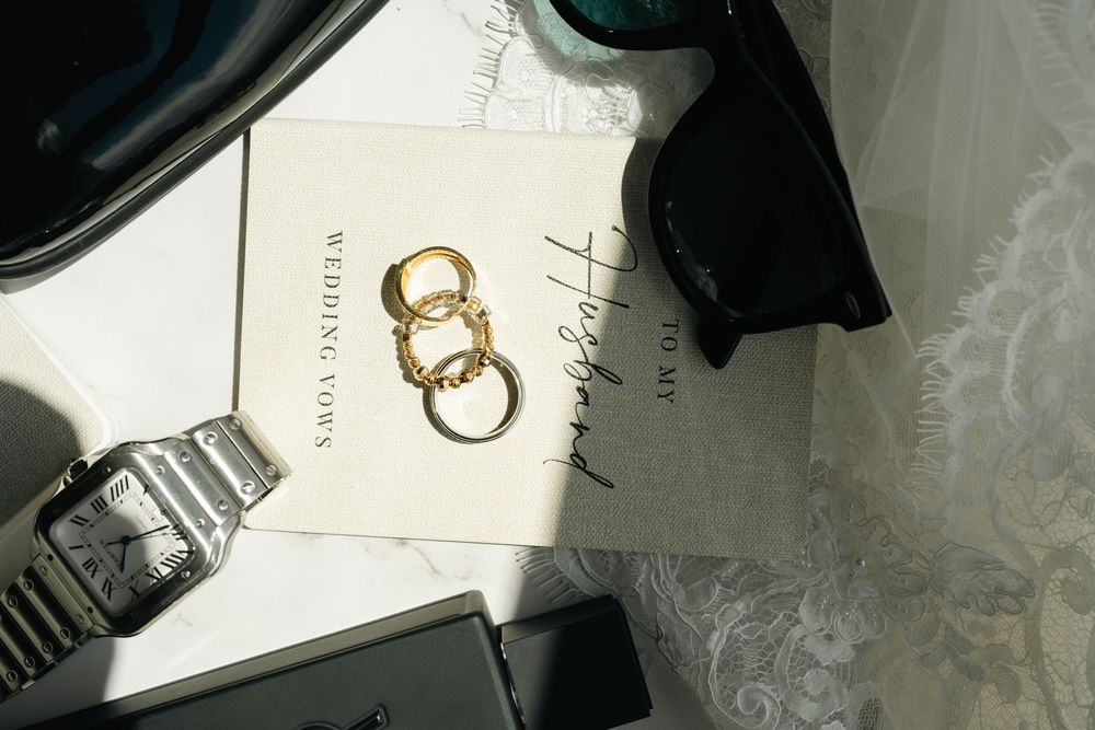 Gold wedding rings on white stationery with sunglasses and watch flatlay detail shot.