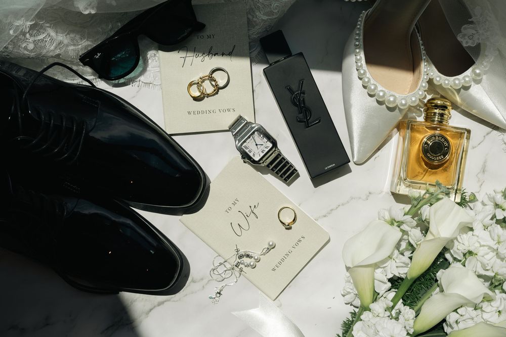 Elegant wedding flat lay with black Oxford shoes, gold rings, pearl heels, YSL perfume, watch, and white floral bouquet.