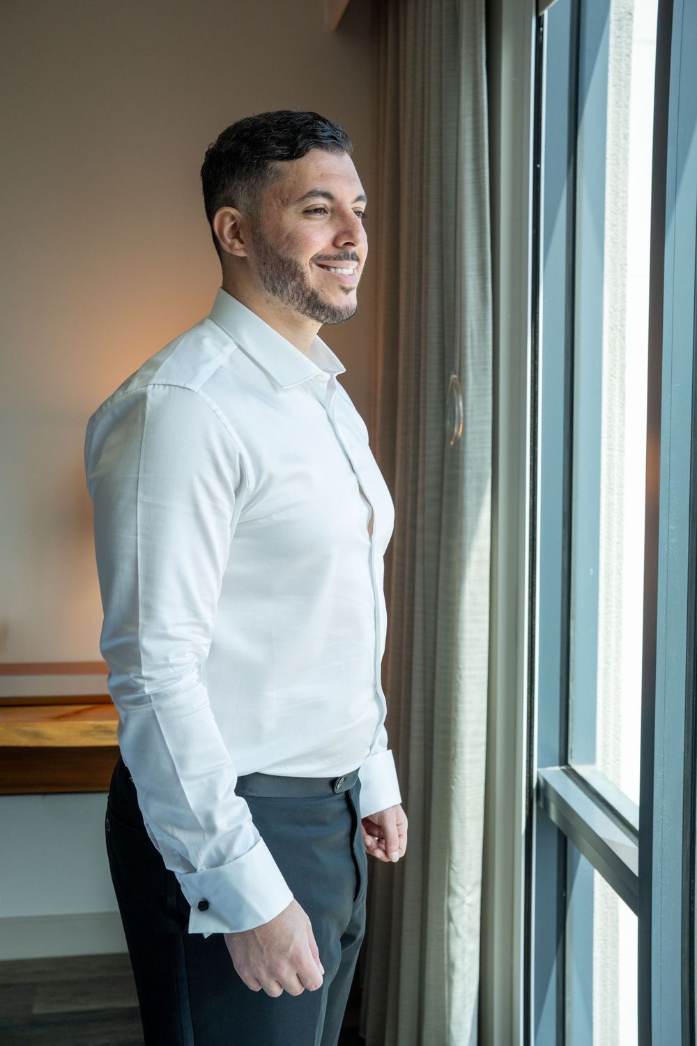 Smiling groom in white dress shirt and grey trousers standing by hotel window on wedding day.