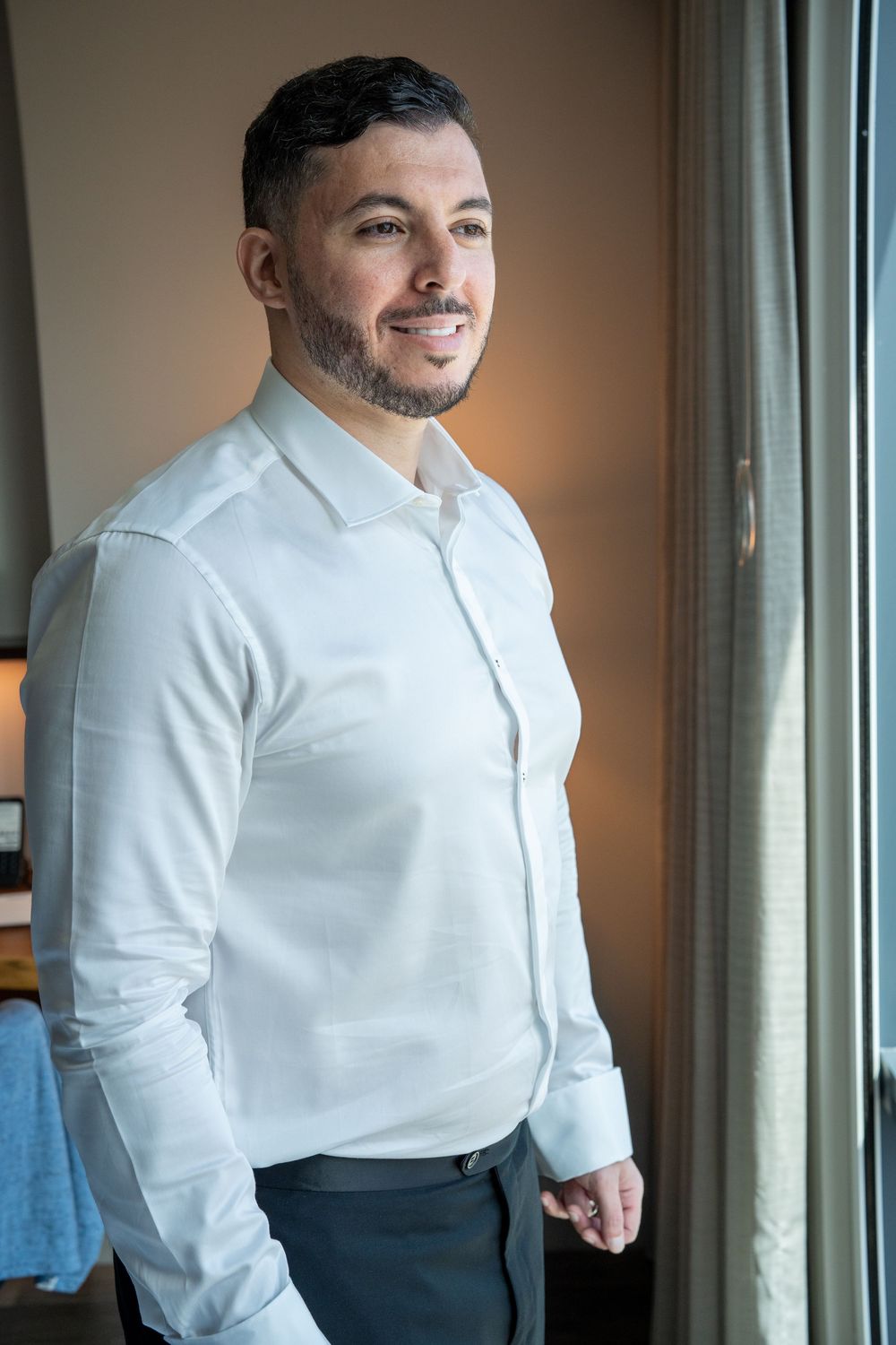 Groom in white dress shirt smiling by window during wedding day preparations.