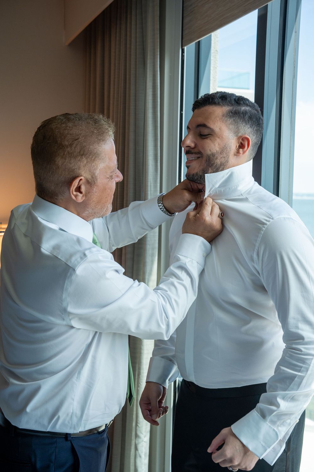 Two men in white dress shirts, one helping the other adjust his collar near a window with ocean views.