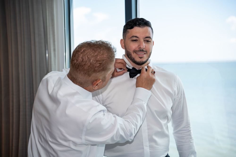 Groom smiling as groomsman helps fasten black bow tie near ocean-view window before wedding.