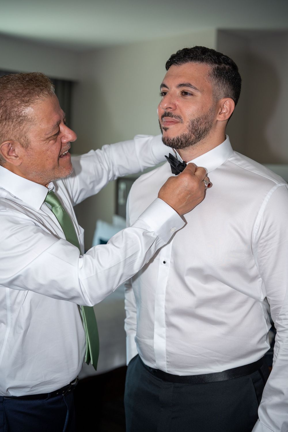 Father helping groom adjust black bow tie while smiling during wedding preparations.
