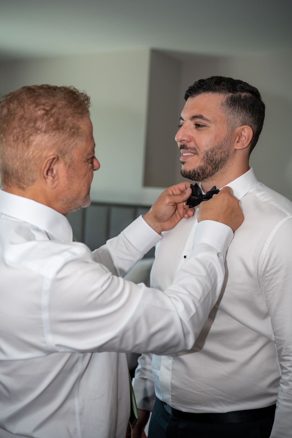 Father and groom share a smile while adjusting bow tie during wedding day getting ready.