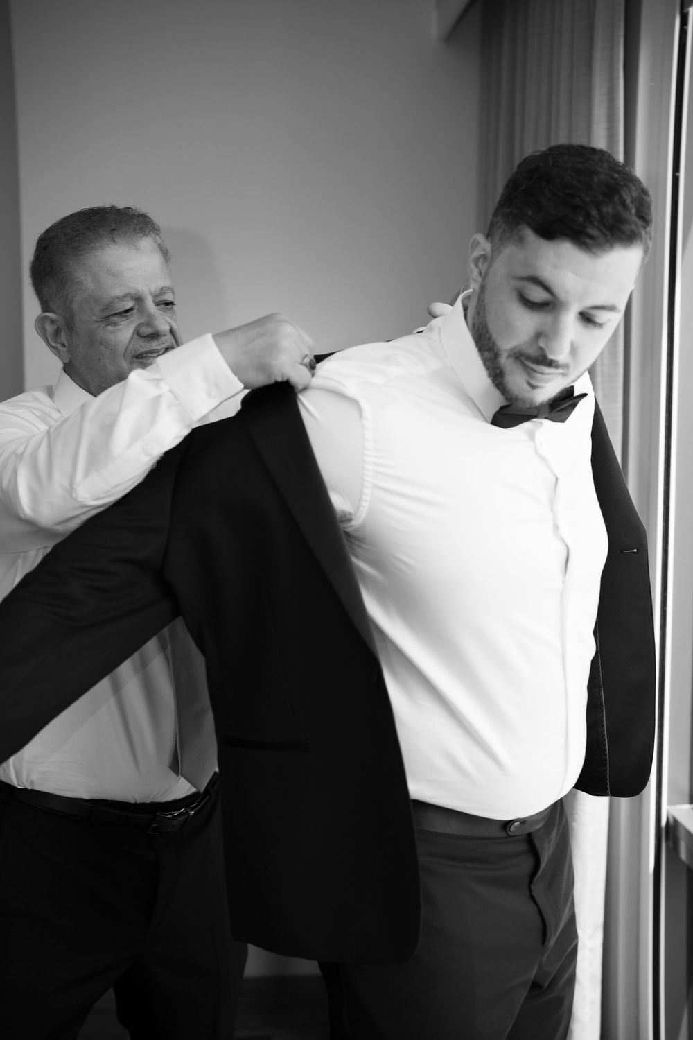 Black and white photo of older man helping groom put on suit jacket before wedding.