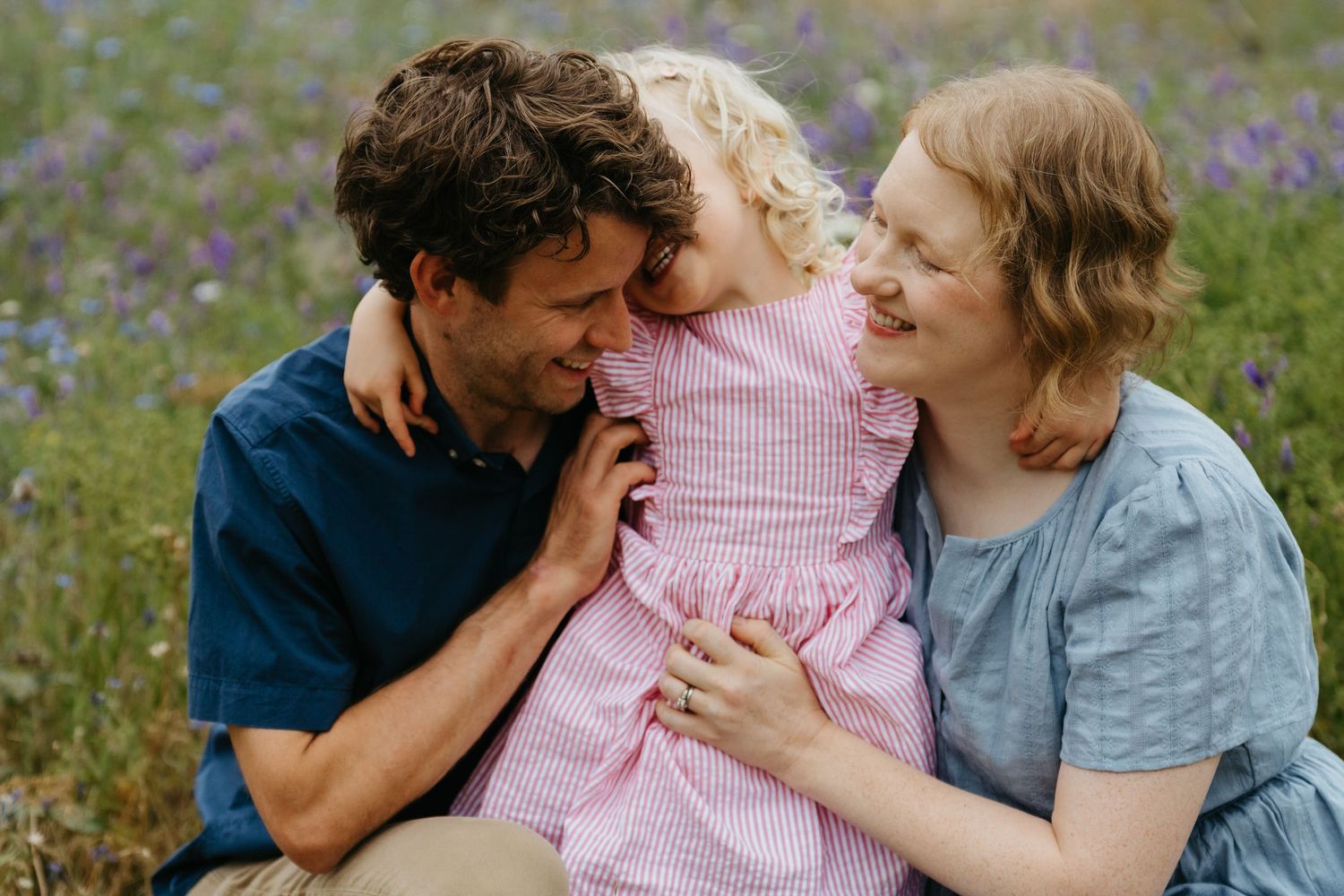 A joyful family of three laughs together in a wildflower field, parents embracing their young blonde daughter in a pink dress.