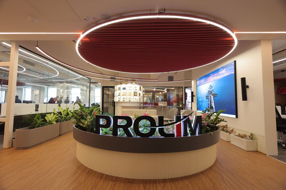 Modern PROLIM corporate office reception area with circular red ceiling light, green plants, and a large digital display screen.