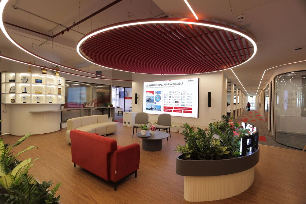 Modern corporate lobby with red circular ceiling light, red sofa, digital display screen, and lush green plants on wood flooring.