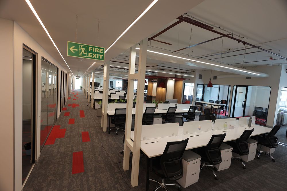Modern open-plan office with white desks, black chairs, fire exit sign, and colorful carpet tiles under bright ceiling lights.