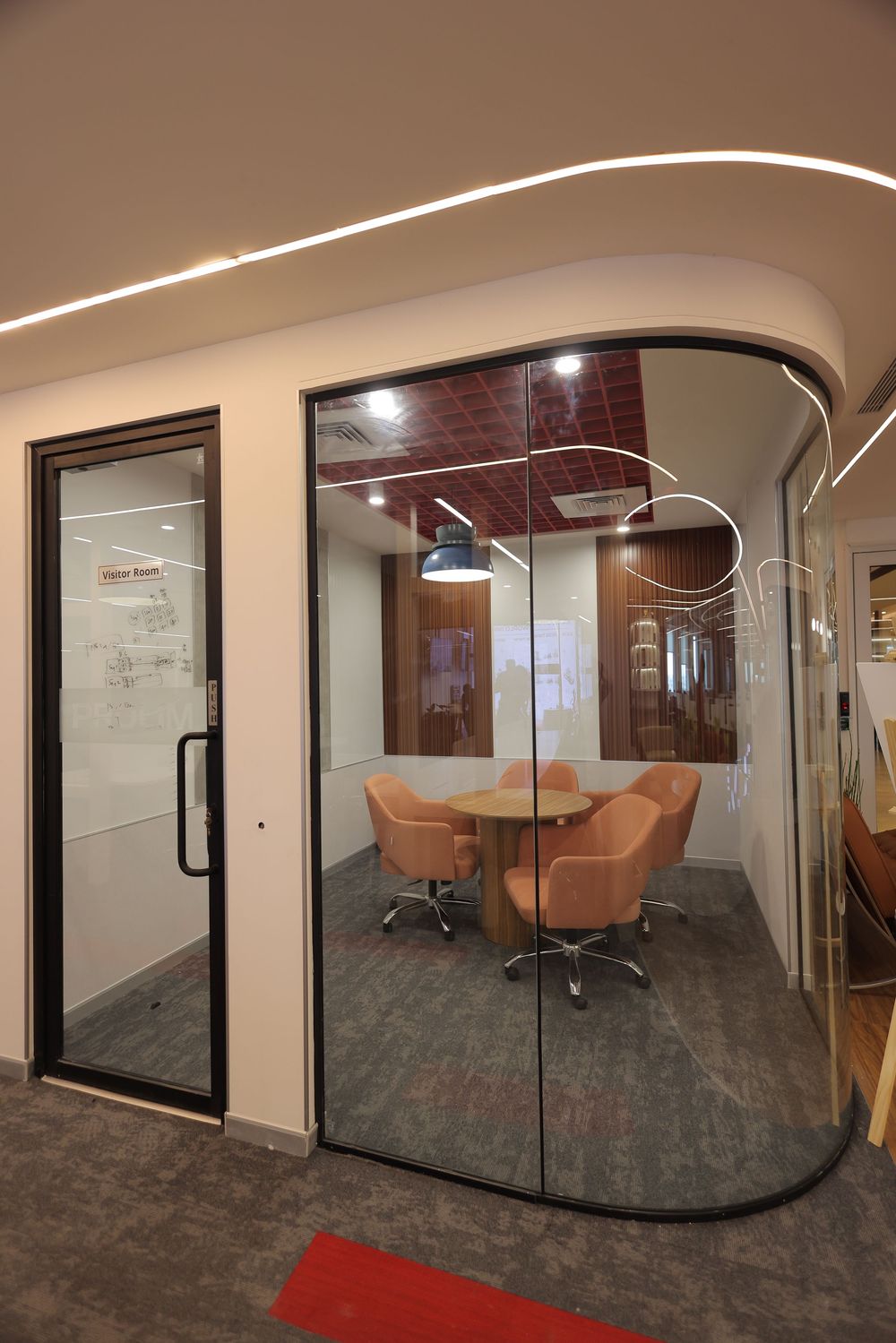 Modern glass-walled office meeting room with curved walls, orange chairs, round table, and elegant LED lighting.