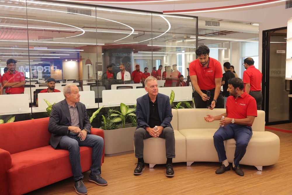 Three professionals in a modern office lounge, two in suits and one in red uniform, engaged in a business discussion.