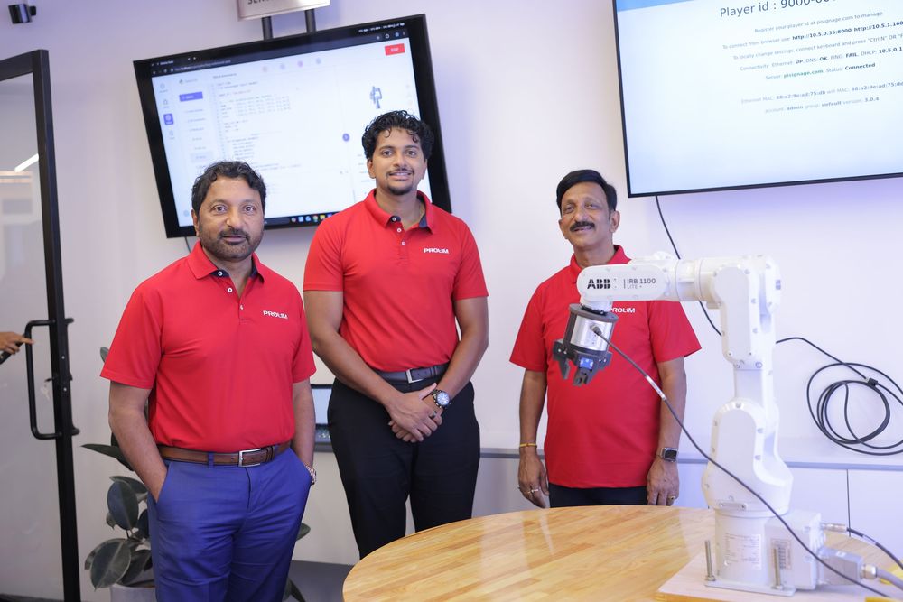 Three men in red ABB polo shirts stand together near a robotic arm display in a modern office setting.