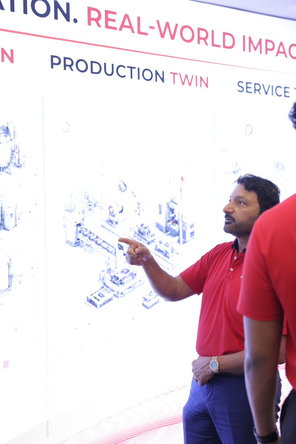 Man in red polo shirt points at a digital production twin display board at a real-world impact technology exhibition.