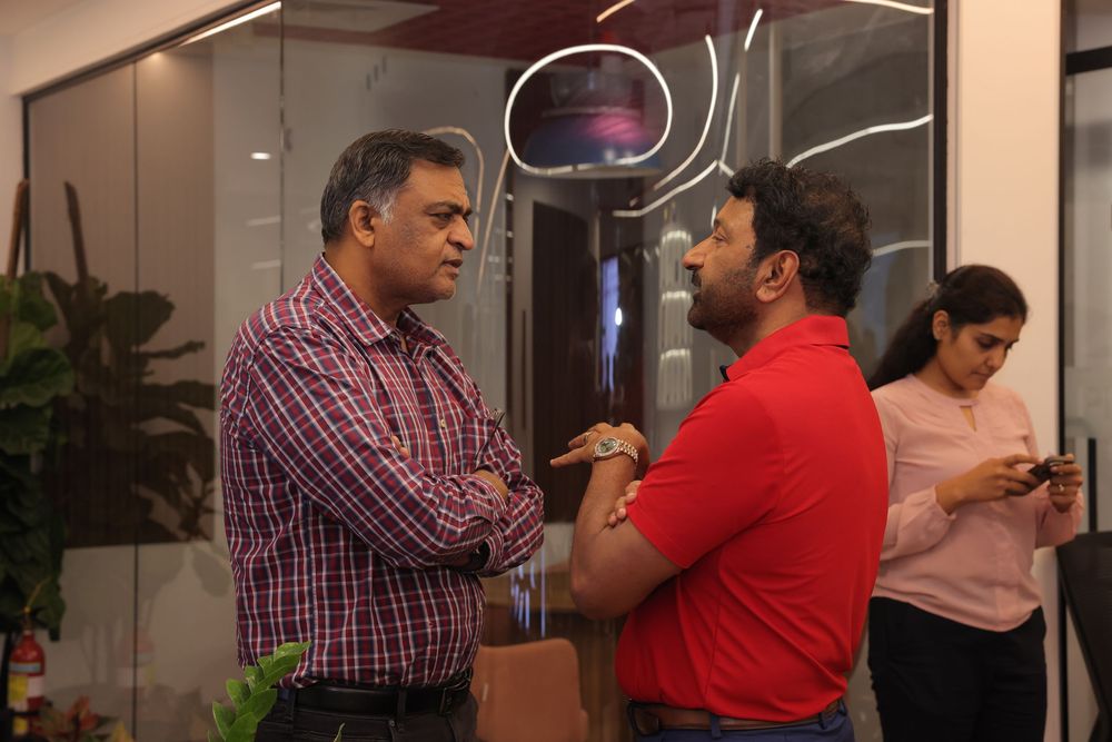 Two men in conversation at an indoor office event, with a woman using her phone in the background.